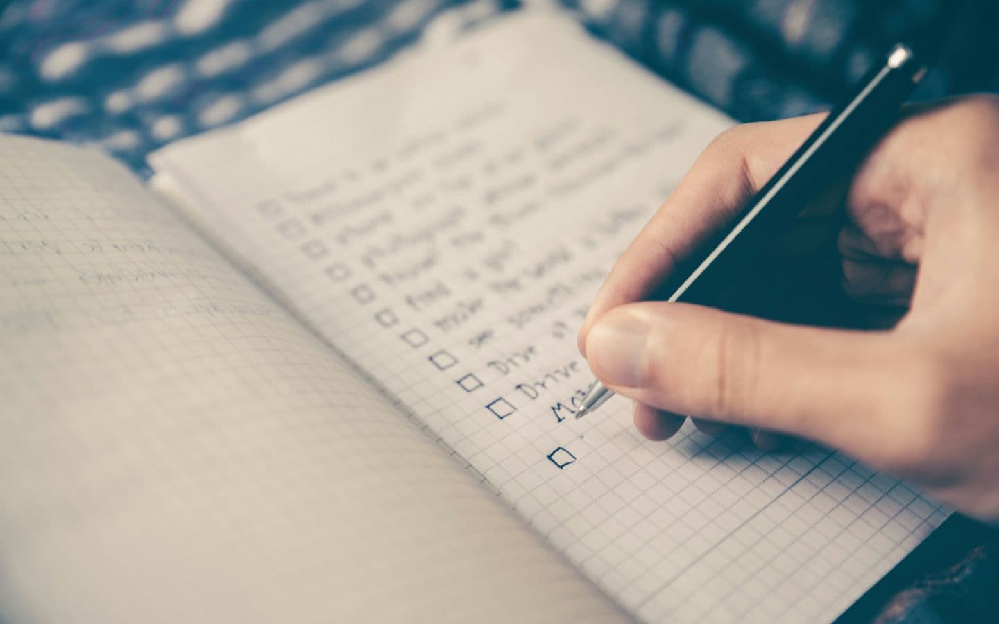 A hand writing a checklist with checkboxes in a grid-lined notebook